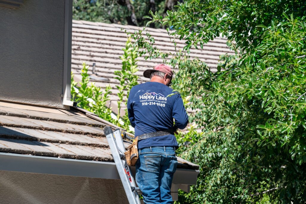 Happy Lites technician installing permanent lights at a roofline beside trees