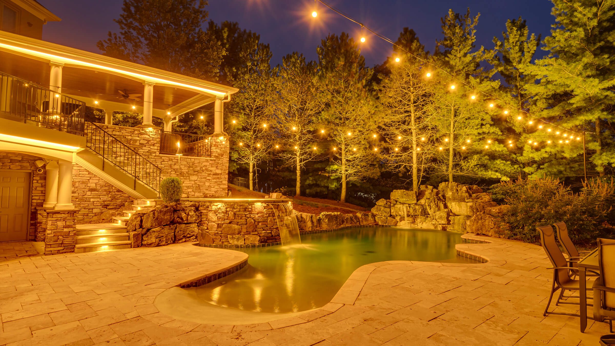 Backyard pool, patio and trees with warm gold permanent outdoor lighting